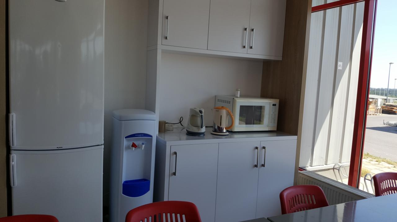 Aneks kuchenny w biurze z białymi szafkami, lodówką, dystrybutorem wody z niebieskim pojemnikiem, czajnikiem elektrycznym, mikrofalówką i czerwonymi krzesłami przy stole, widok z okna na plac...