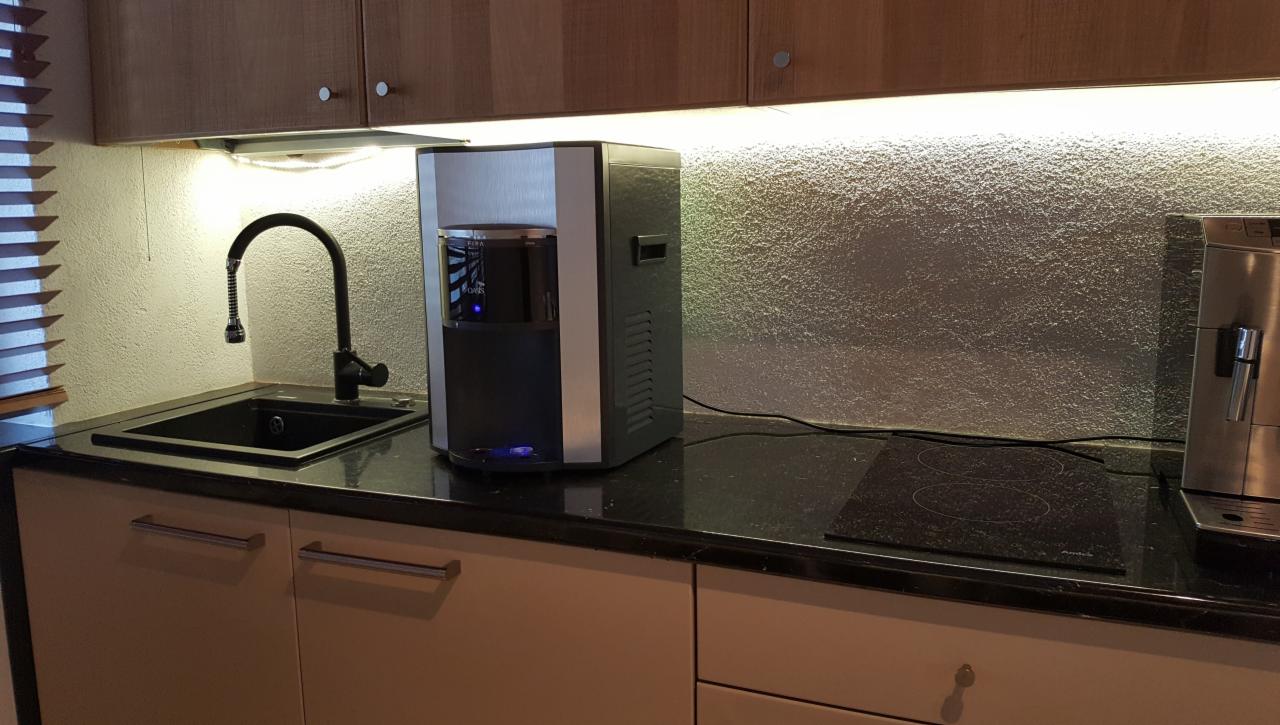 Nowoczesny dystrybutor wody w srebrno-czarnej obudowie umieszczony na blacie kuchennym obok zlewu z czarną baterią i ekspresu do kawy, oświetlony światłem LED z szafek wiszących.