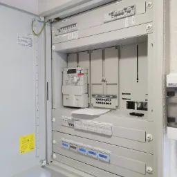 Otwarta szara skrzynka rozdzielcza z licznikiem energii elektrycznej, bezpiecznikami i miernikami, widoczne przewody uziemiające i oznaczenia na etykietach.