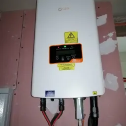 Biały inwerter fotowoltaiczny marki Solis zamontowany na ścianie wykończonej płytami gipsowo-kartonowymi, widoczne kable i złącza, naklejki ostrzegawcze oraz panel sterowania z wyświetlaczem.