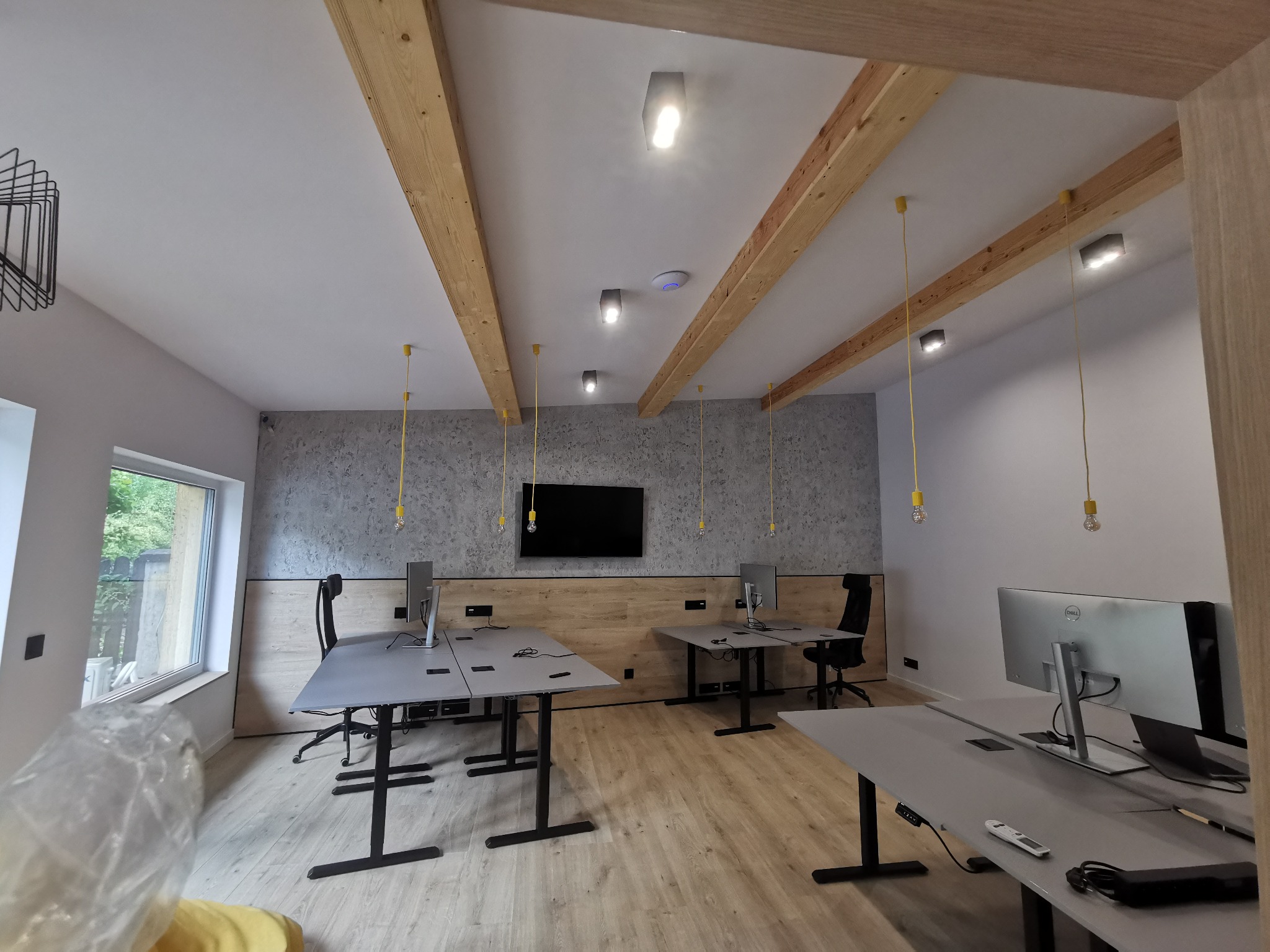Nowoczesne biuro z drewnianymi belkami sufitowymi, szarymi biurkami z czarnymi nogami, wiszącymi lampami z żółtymi kablami i betonową ścianą akcentującą.