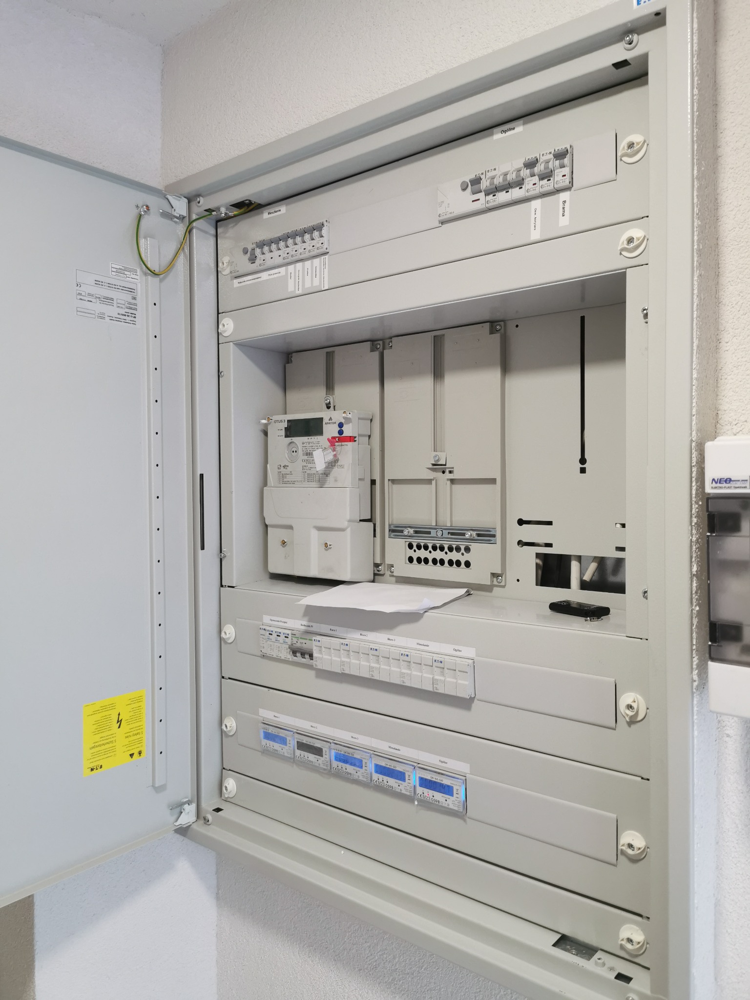 Otwarta szara skrzynka rozdzielcza z licznikiem energii elektrycznej, bezpiecznikami i miernikami, widoczne przewody uziemiające i oznaczenia na etykietach.