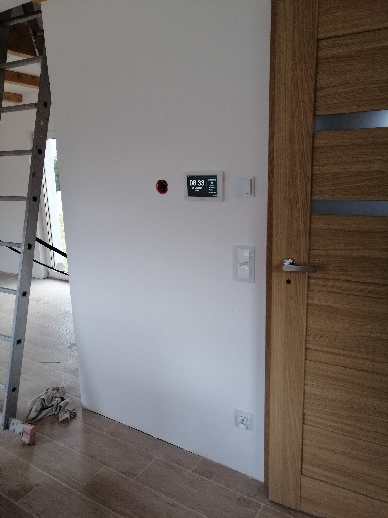 Panel sterowania inteligentnego domu zamontowany na białej ścianie obok drewnianych drzwi z metalową klamką, widoczna drabina i fragment podłogi z płytek.