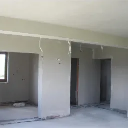 Wnętrze po tynkowaniu maszynowym: gładkie ściany, widoczne kable, surowy stan wykończenia przed malowaniem. Okno z widokiem na zieleń.