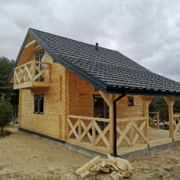 Surowy, drewniany dom z bali z balkonem i zadaszonym tarasem, położony na piaszczystym terenie, z widocznym stosem drewna opałowego.