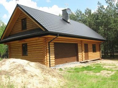 Drewniany dom z bali z dwuspadowym dachem pokrytym ciemną dachówką, widoczny komin z anteną satelitarną, zamknięte brązowe drzwi garażowe, na pierwszym planie pryzma piasku.