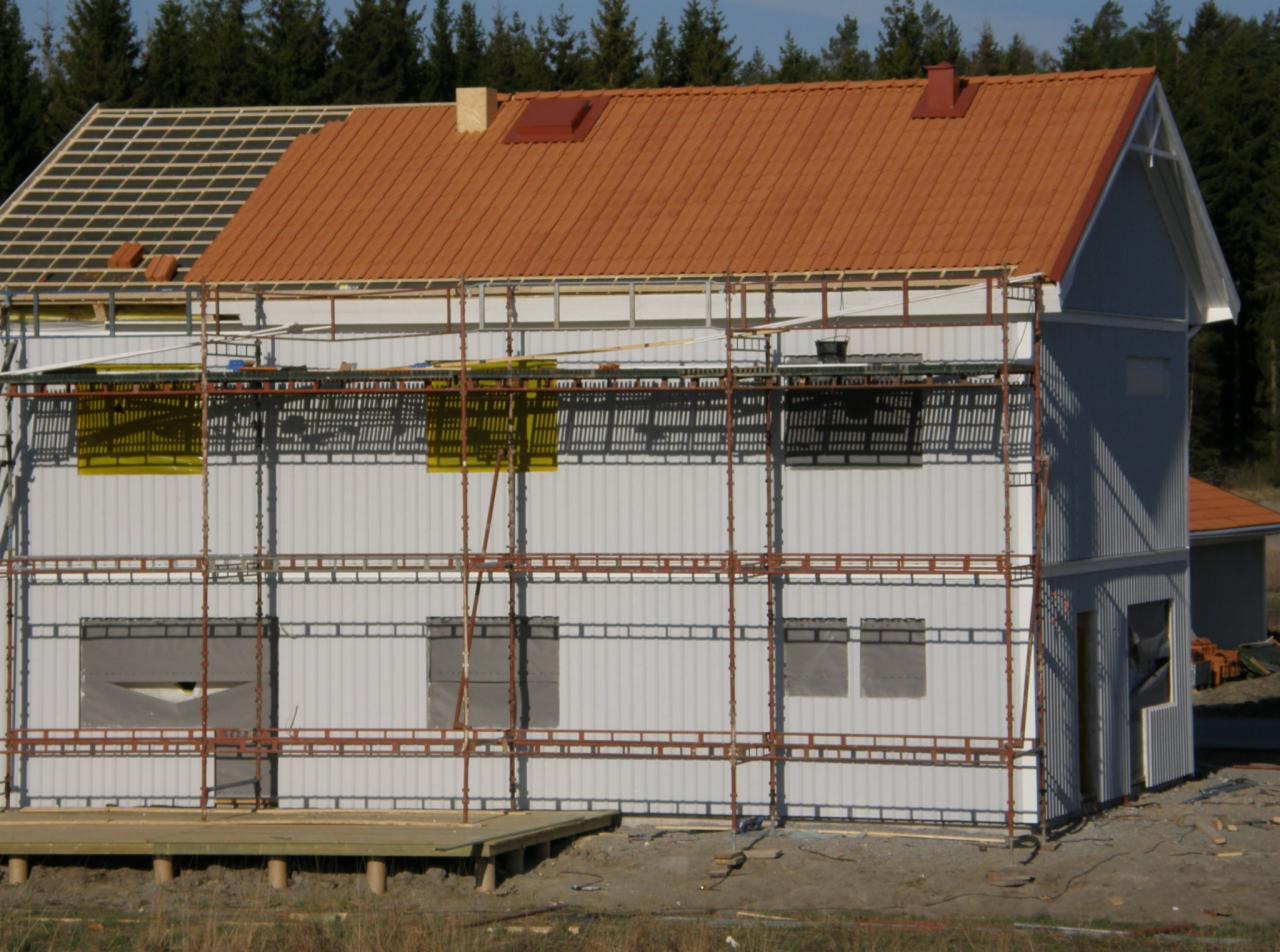 Wznoszenie dwukondygnacyjnego domu szkieletowego z częściowo ułożonym dachem z czerwonej dachówki. Widoczne rusztowania i niezabudowane okna. W tle las iglasty.