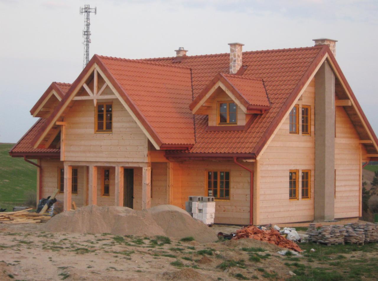 Surowy dom szkieletowy z czerwoną dachówką, widoczne materiały budowlane na pierwszym planie, w tle maszt telekomunikacyjny.