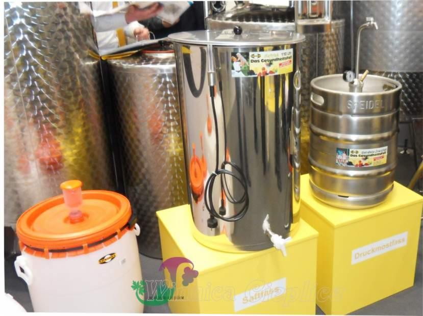 Różne typy zbiorników ze stali nierdzewnej i tworzyw sztucznych do przechowywania wina i miodu, umieszczone na żółtych podstawkach, w tle metalowe zbiorniki o teksturowanej powierzchni.