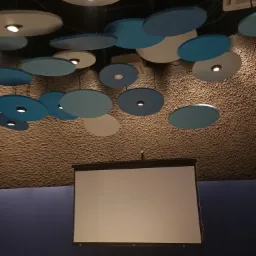 Nowoczesny sufit z okrągłymi panelami akustycznymi w odcieniach bieli i błękitu, oświetlenie punktowe, z widocznym ekranem projekcyjnym w pomieszczeniu biurowym.