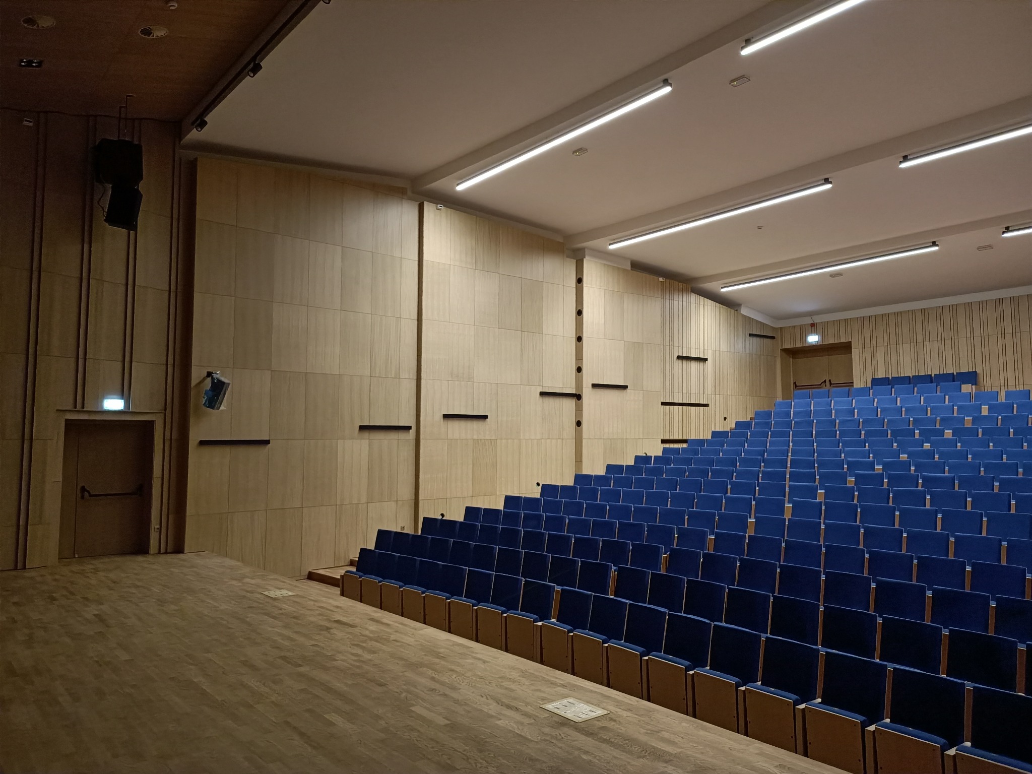 Wnętrze sali audytoryjnej z rzędami niebieskich krzeseł, drewnianą podłogą i ścianami wyłożonymi panelami w odcieniach beżu, oświetlonej przez podłużne lampy sufitowe.