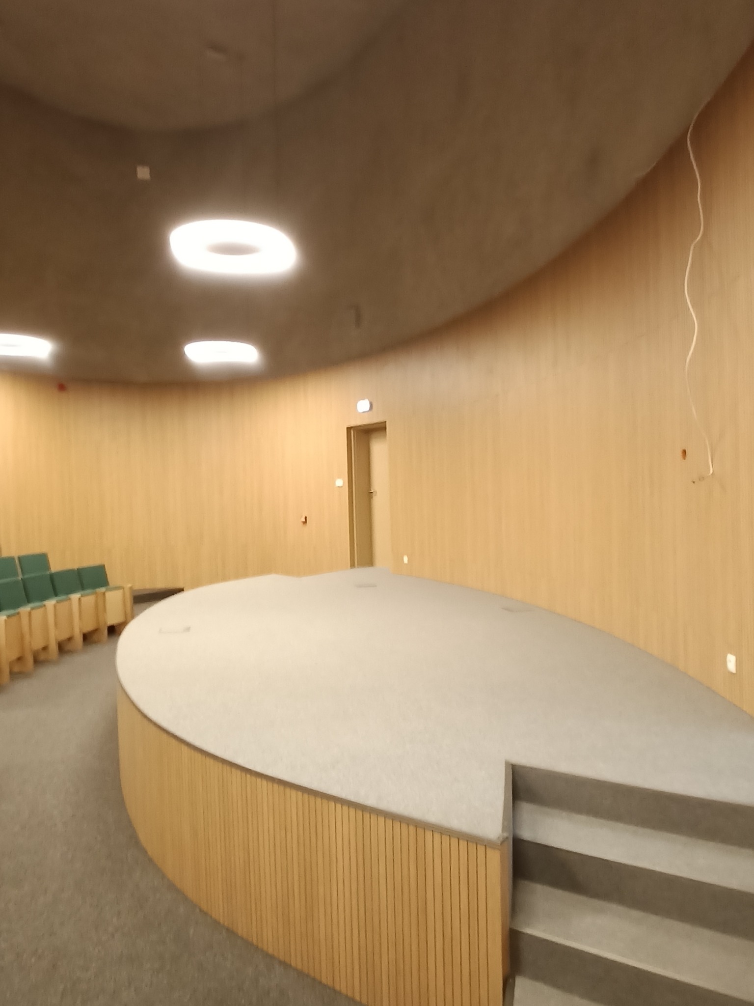 Wnętrze sali konferencyjnej z drewnianymi panelami ściennymi, podwyższoną sceną z trzema stopniami i rzędem zielonych krzeseł w tle.