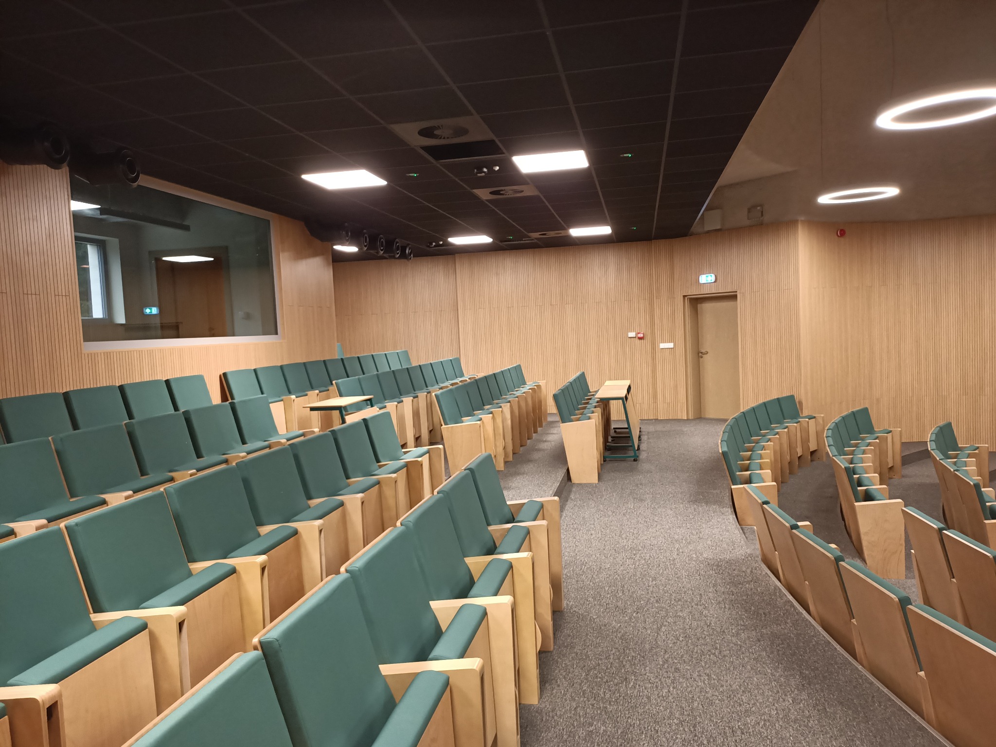 Sala konferencyjna z rzędami zielonych krzeseł i drewnianymi ścianami, oświetlona panelami LED i okrągłymi lampami sufitowymi.