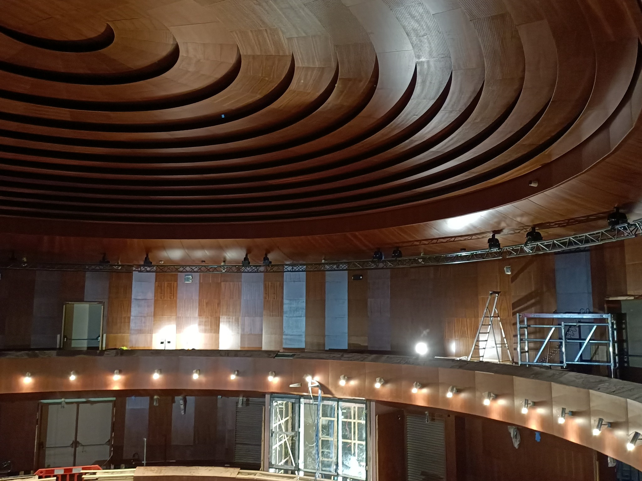 Wnętrze budynku w trakcie wykończenia, widoczna konstrukcja sufitu z drewnianymi panelami ułożonymi warstwowo, ściany z fragmentami płyt gipsowo-kartonowych, oświetlenie robocze i drabina.