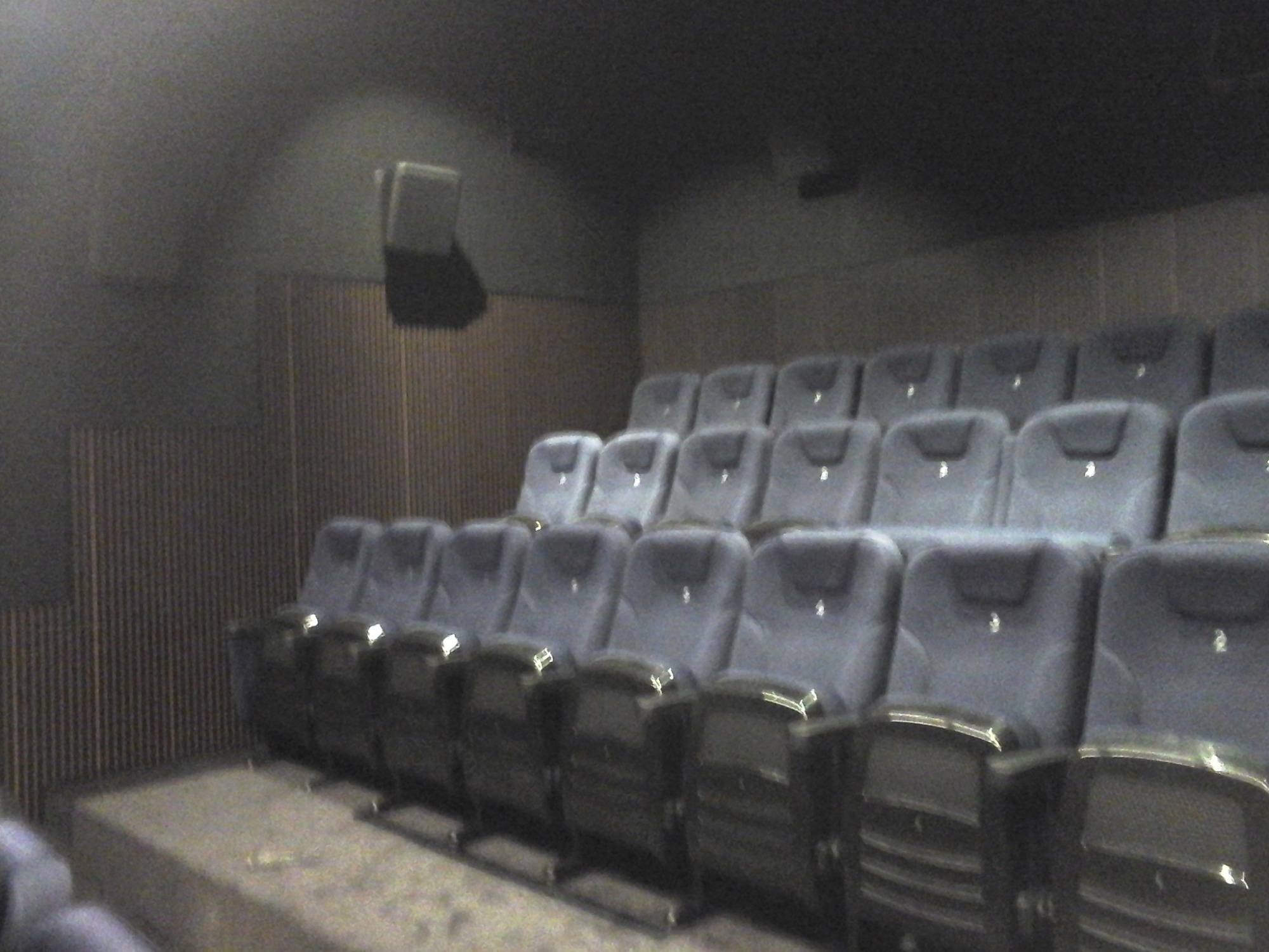 Rzędy pustych, niebieskich foteli w zaciemnionej sali kinowej z widocznym głośnikiem na ścianie.