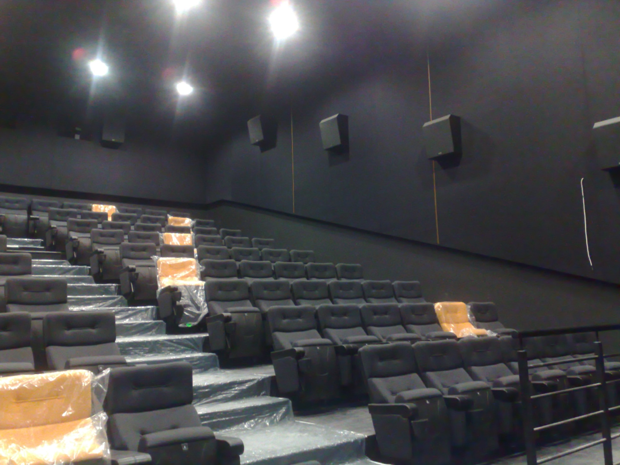 Wnętrze sali kinowej z rzędami czarnych foteli częściowo zabezpieczonych folią ochronną, widoczne czarne ściany i głośniki, oświetlenie sufitowe.