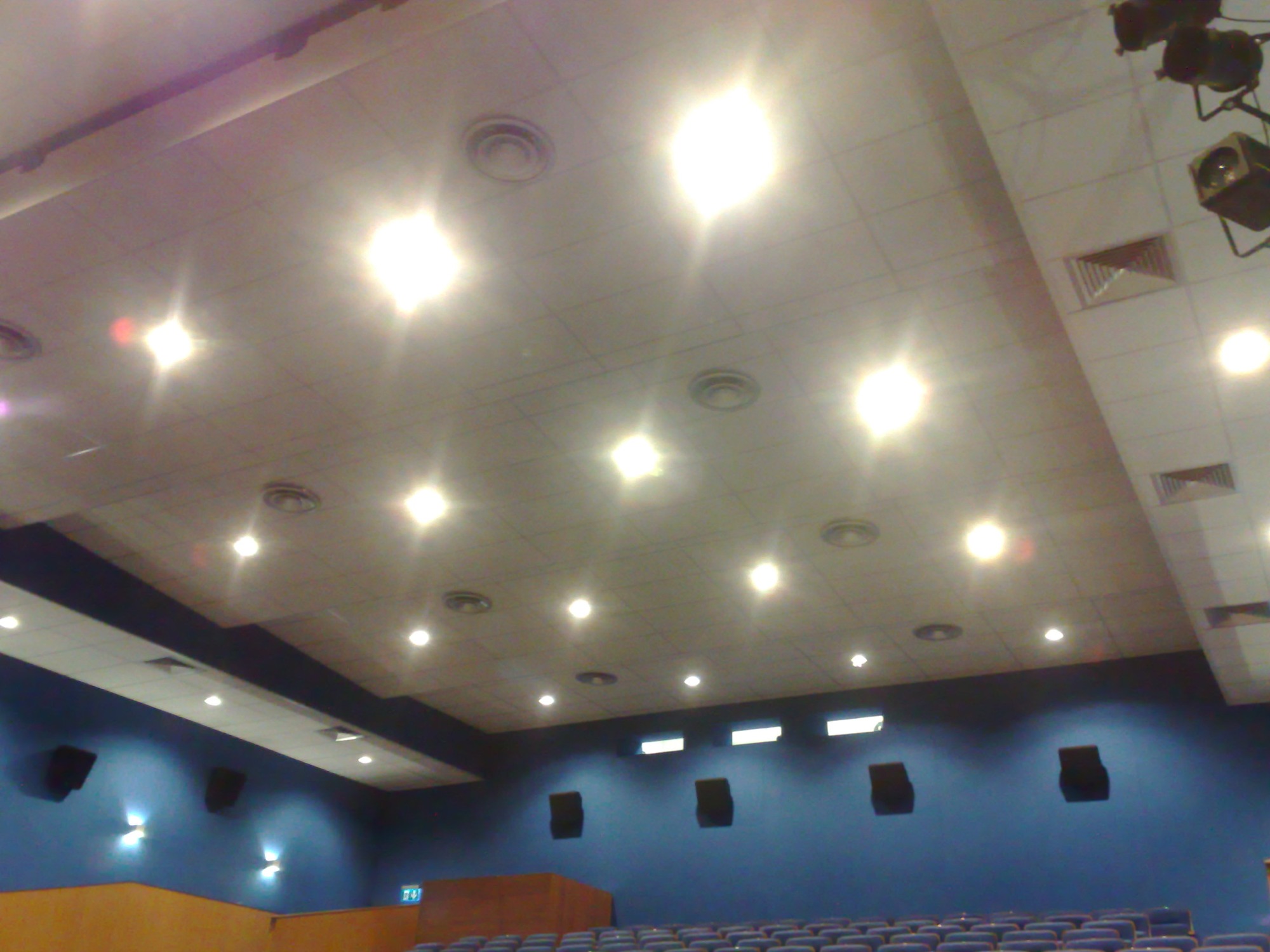 Wnętrze sali kinowej z widocznym sufitem podwieszanym z oświetleniem punktowym i kratkami wentylacyjnymi, ciemnoniebieskie ściany z zamontowanymi głośnikami oraz rzędy foteli kinowych w dolnej...