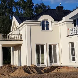 Nowoczesny dom jednorodzinny z jasną elewacją, balkonami z balustradami i dużymi oknami, teren wokół domu częściowo rozkopany.