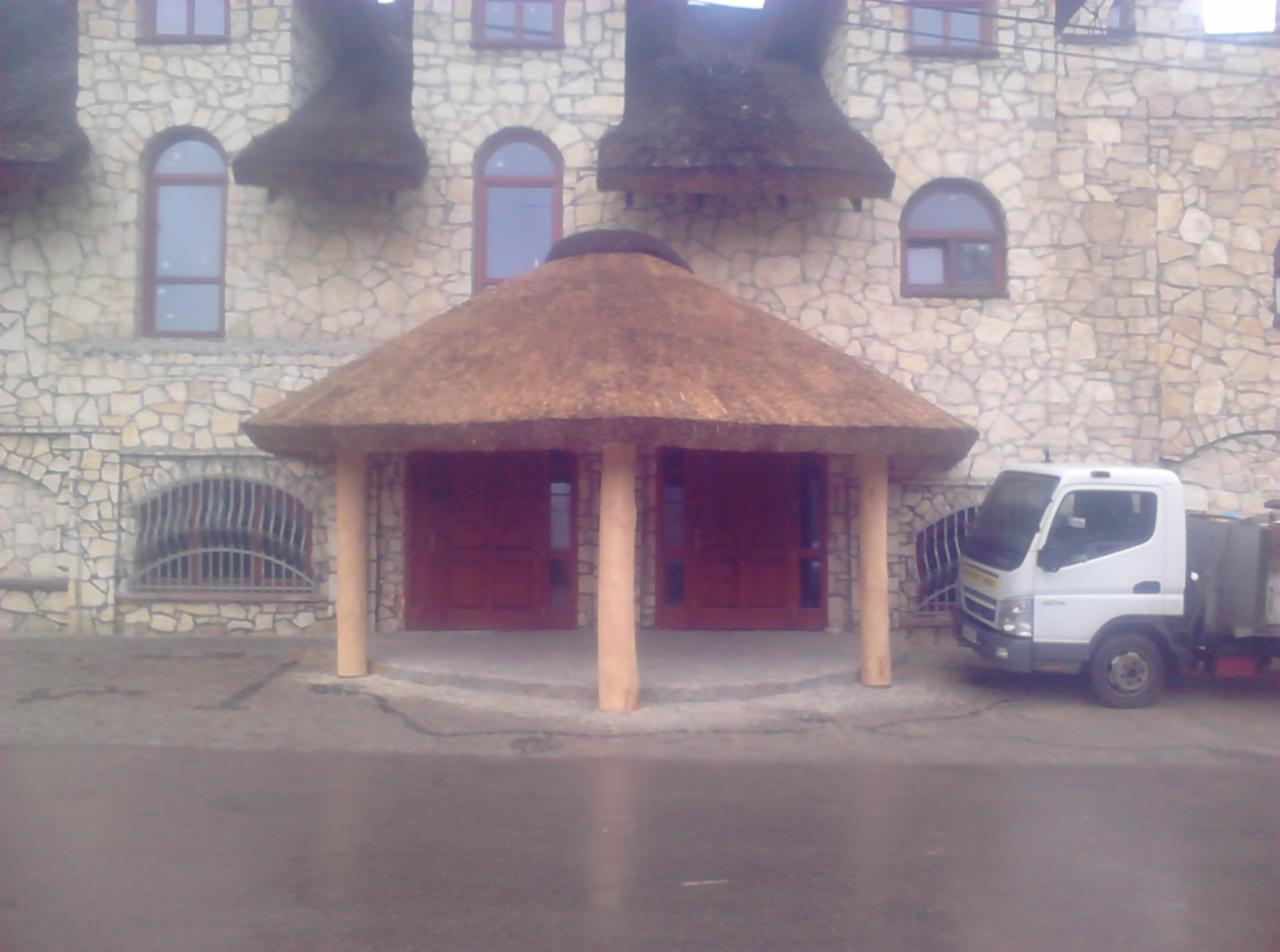 Kamienny budynek z wejściem zadaszonym strzechą trzcinową, wspartym na drewnianych kolumnach, obok zaparkowany biały samochód dostawczy.