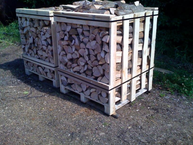 Dwa kontenery paletowe wypełnione pociętym drewnem kominkowym, zabezpieczone czarnymi taśmami, stojące na żwirowym podłożu z zielenią w tle.