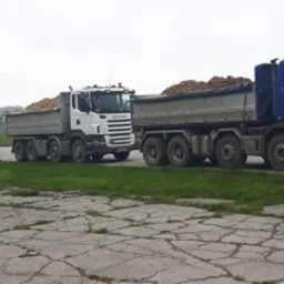 Trzy ciężarówki wywrotki (niebieska, biała i niebieska) ustawione jedna za drugą na betonowym placu z zielenią po bokach. Niebieska ciężarówka z przodu załadowana ziemią.