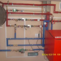 Złożona instalacja centralnego ogrzewania z czerwonymi rurami izolowanymi, niebieskimi rurami i dużym zbiornikiem akumulacyjnym, widoczne zawory, manometry i zabezpieczenia, zamontowana na beżowej...