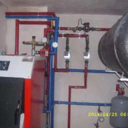 Kotłownia z kotłem marki Fitz, zasobnikiem ciepłej wody i rozbudowaną instalacją hydrauliczną w kolorach czerwonym i niebieskim, manometry i zawory na ścianie.