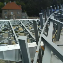 Wznoszenie stalowej konstrukcji z krzyżulcami na betonowym fundamencie, widok z góry, w tle budynek mieszkalny i drzewa.