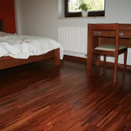 Ciemna, lakierowana podłoga parkietowa ułożona w jodełkę w sypialni z drewnianym łóżkiem i biurkiem przy oknie.