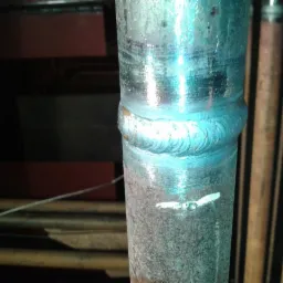 Metalowa rura połączona spoiną z charakterystycznym niebieskim zabarwieniem. Widoczne ślady obróbki i spawania. W tle inne rury i elementy konstrukcyjne.