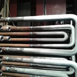 Rzędy rur kotłowych, niektóre z widocznymi spawami, inne pokryte szarą farbą. Na rurach numery identyfikacyjne. Widoczne spoiny i złącza.