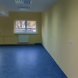 Pomalowane na żółto wnętrze pomieszczenia z niebieską podłogą, grzejnikami pod oknami z kratami i widocznymi puszkami elektrycznymi na ścianie.