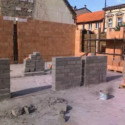 Wznoszenie ścian z bloczków ceramicznych Porotherm na budowie domu jednorodzinnego, widoczne zbrojenie pionowe i stos rusztowań w tle.