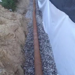 Drenaż opaskowy budynku w trakcie wykonywania: rura drenarska obsypana żwirem, zabezpieczona geowłókniną, widoczny fragment ściany fundamentowej z izolacją.