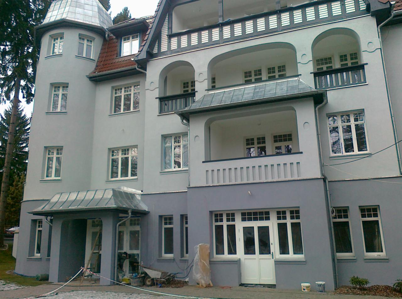 Dwukondygnacyjny dom z wieżyczką i balkonami, świeżo po malowaniu elewacji na odcienie szarości, z widocznymi narzędziami i materiałami budowlanymi sugerującymi trwające prace wykończeniowe.