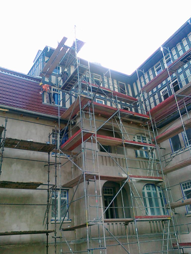 Prace elewacyjne na budynku z ceglanym dachem, widoczne rusztowania i pracownicy w odzieży roboczej, okna zabezpieczone folią.