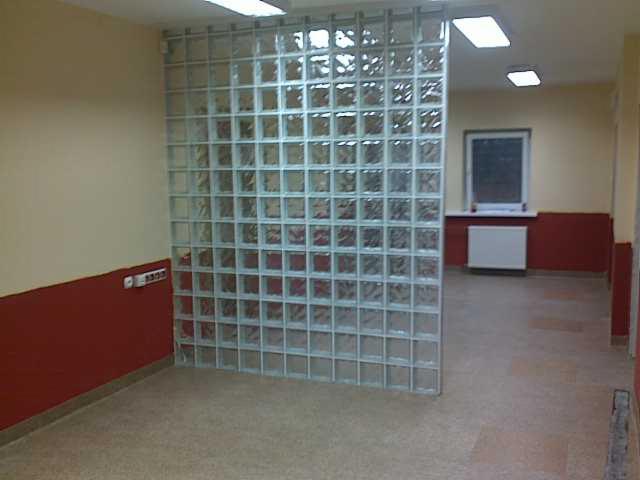 Ścianka działowa z luksferów w surowym wnętrzu biurowym z czerwonymi i beżowymi ścianami, widoczne okno i grzejnik.