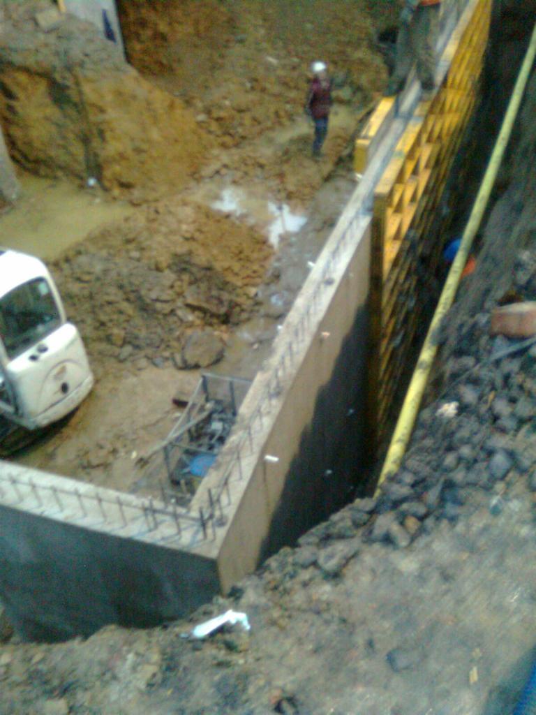 Budowa fundamentów w wykopie z użyciem szalunków, widoczny pracownik w oddali i maszyna budowlana.