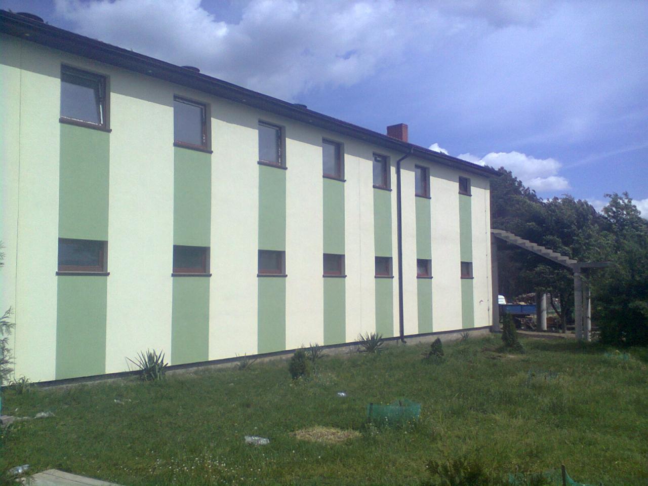 Budynek z elewacją w pionowe pasy w odcieniach zieleni i bieli, widoczne okna z brązowymi ramami, trawnik i fragment konstrukcji tarasu w tle.