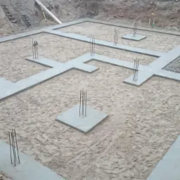 Wylany fundament z betonu z wystającymi prętami zbrojeniowymi na piaszczystym podłożu w wykopie, widoczne ślady stóp i kałuża wody w rogu.