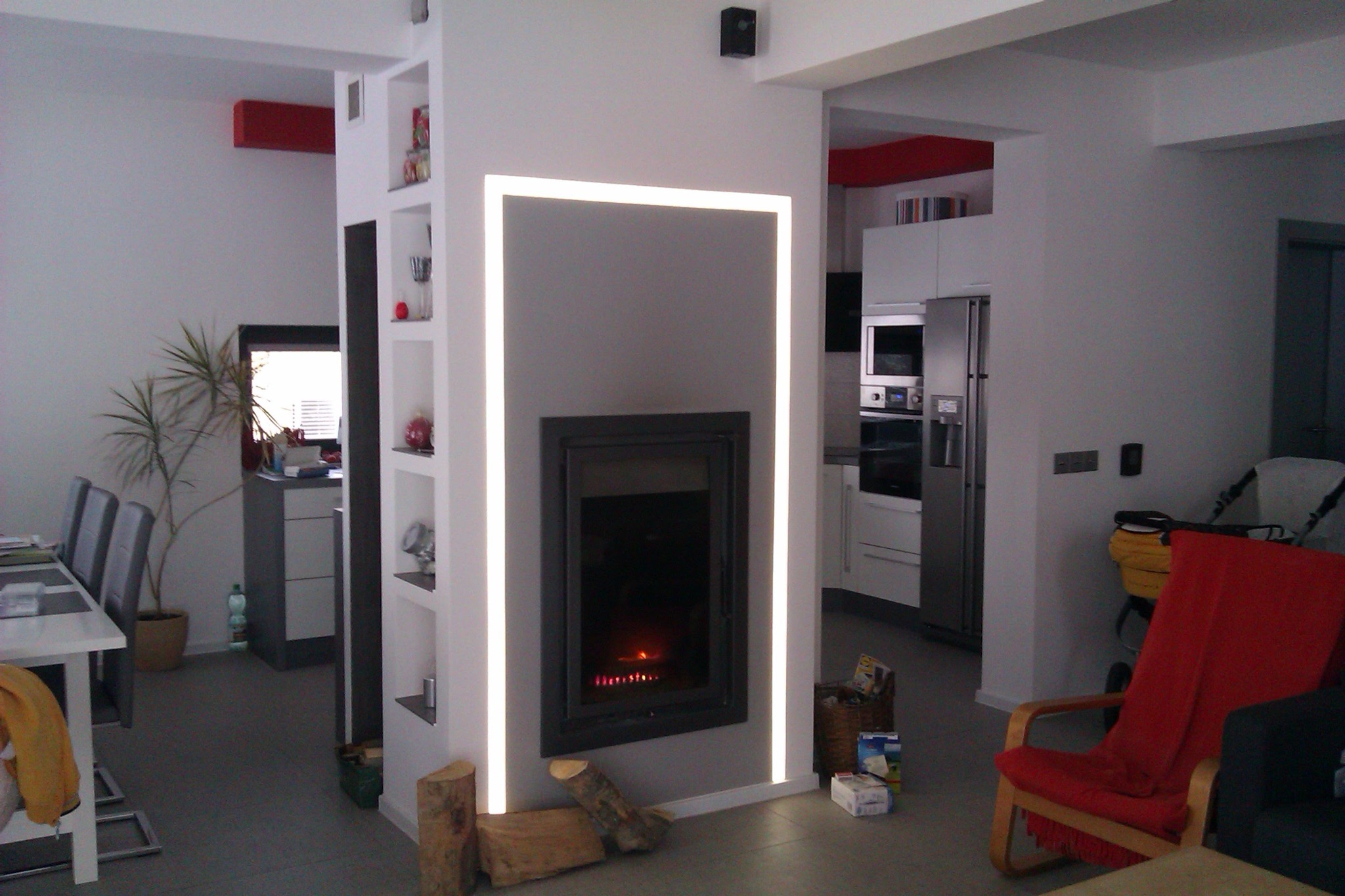 Nowoczesny salon z kominkiem obudowanym w szarą ścianę z podświetleniem LED, widoczne drewno do kominka i fragment kuchni w tle.