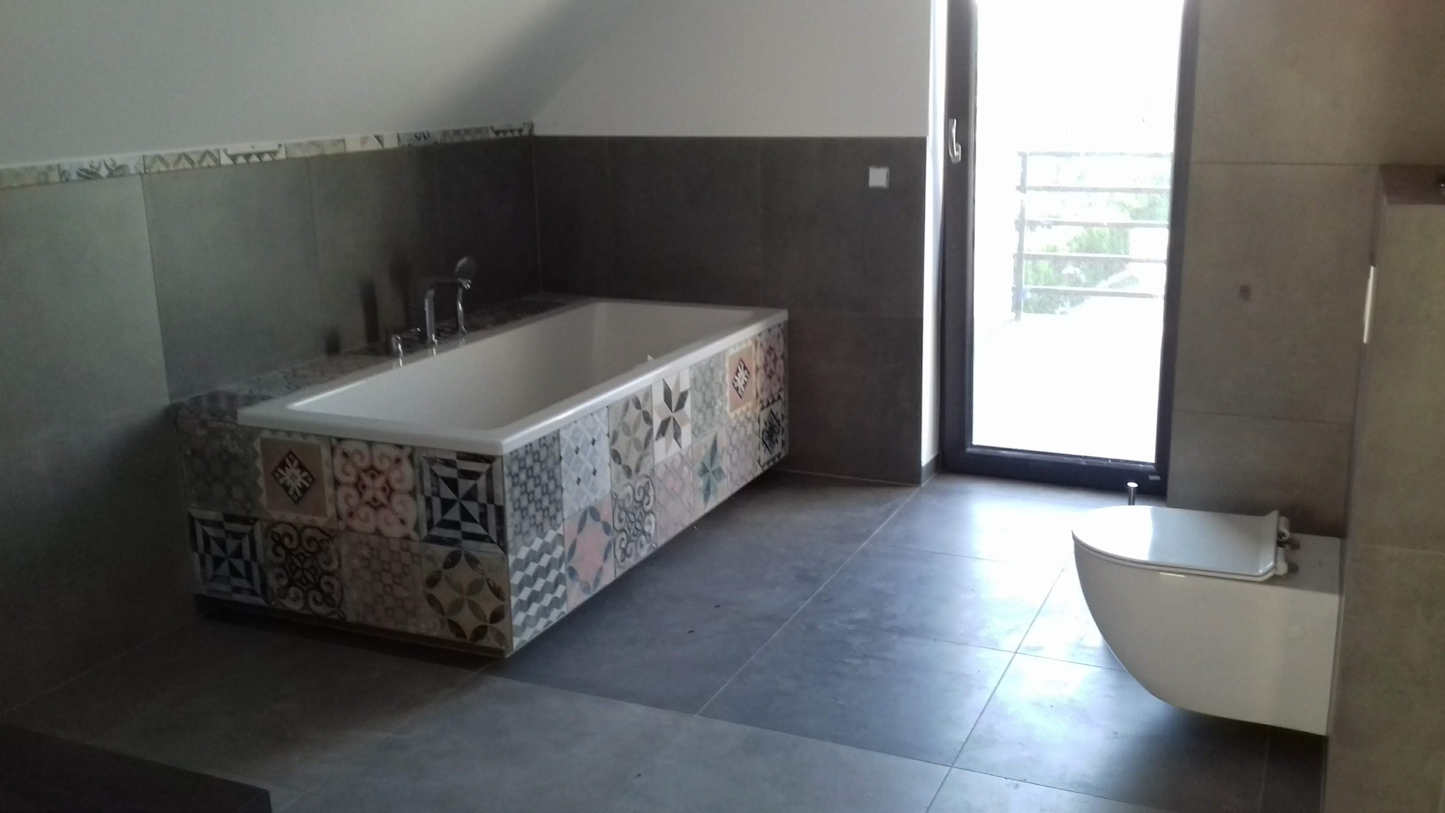 Nowoczesna łazienka na poddaszu z wanną obudowaną dekoracyjnymi płytkami, wiszącą toaletą i dużym oknem.