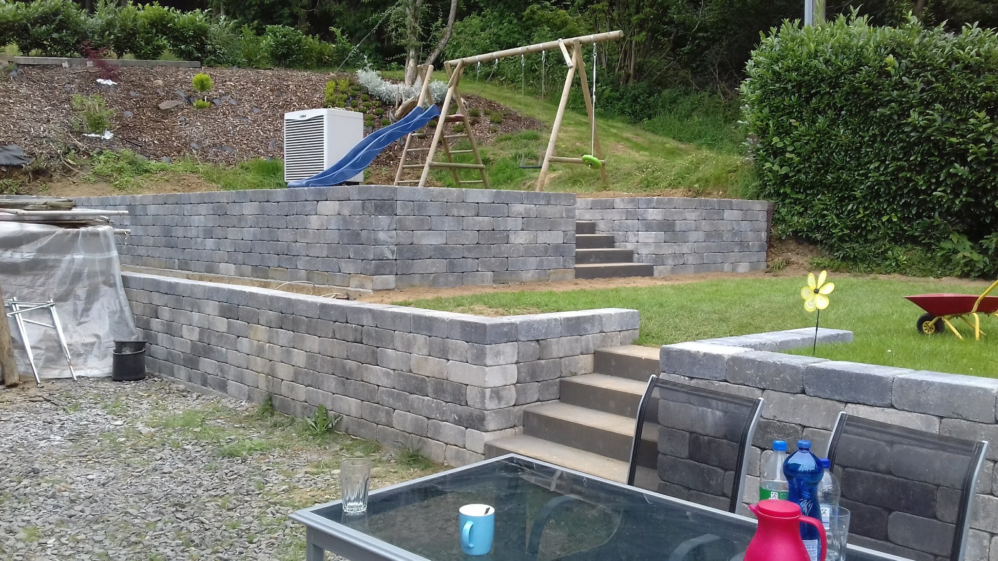 Murowana, dwupoziomowa ściana oporowa z szarej kostki w ogrodzie w Dobczycach, z widocznymi schodami, huśtawką i zjeżdżalnią w tle.