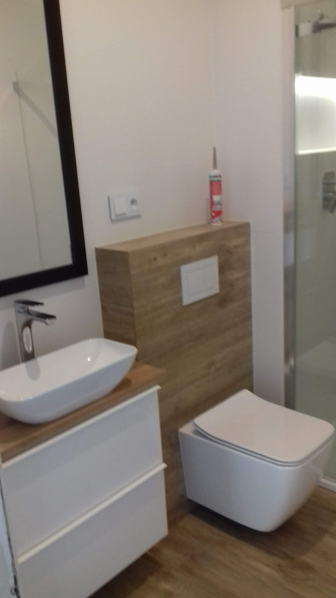 Nowoczesna łazienka z podwieszaną miską WC, umywalką nablatową na szafce z szufladami, lustrem w czarnej ramie i kabiną prysznicową; widoczna tuba silikonu na obudowie stelaża WC.