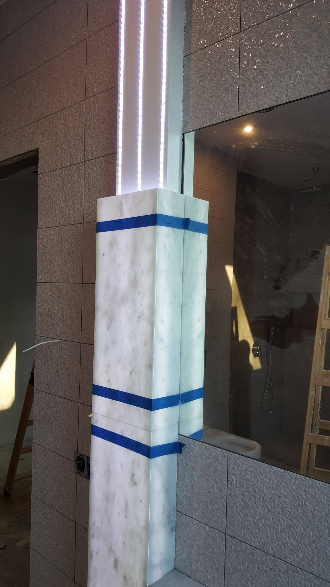 Wykończona kolumna z podświetleniem LED w łazience, zabezpieczona niebieską taśmą malarską, odbicie w lustrze ukazuje fragment kabiny prysznicowej i drabiny.