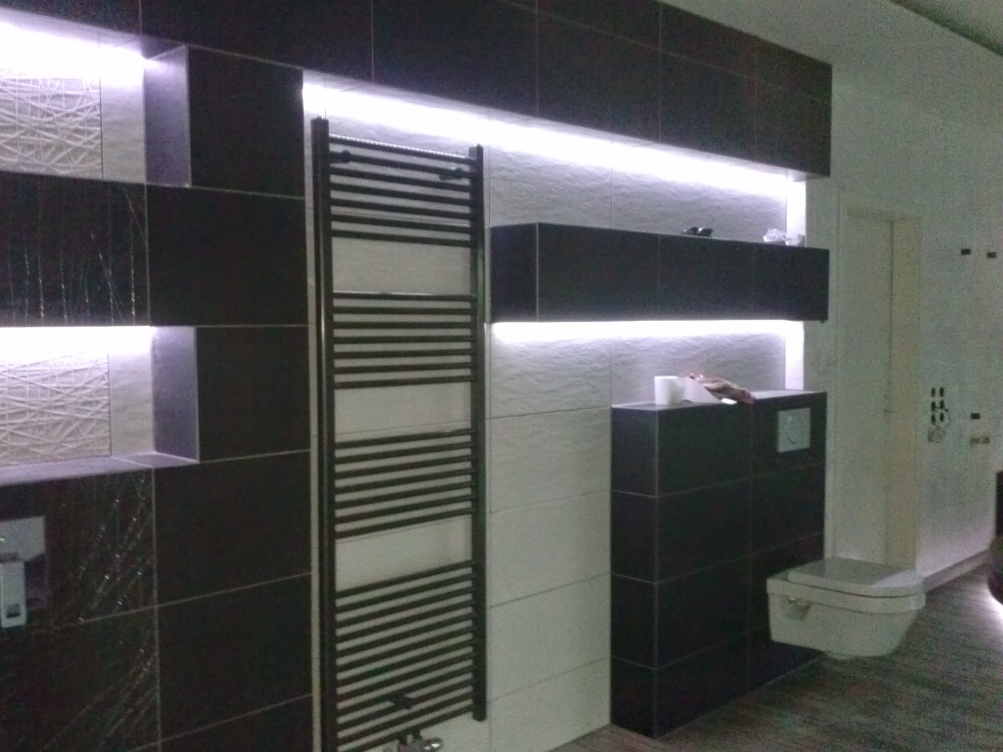 Nowoczesna łazienka z czarno-białymi płytkami, podświetleniem LED, grzejnikiem drabinkowym i podwieszaną toaletą.