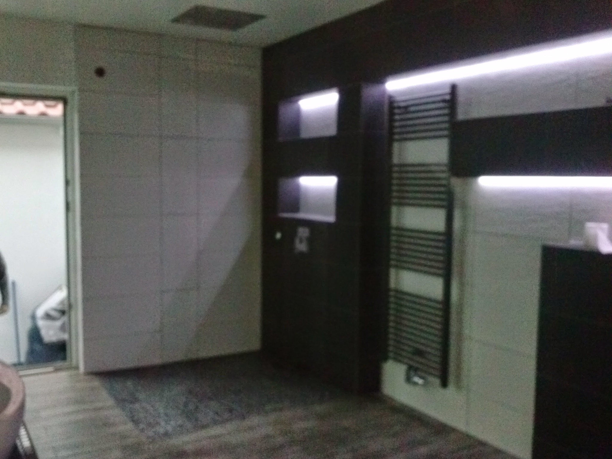 Nowoczesna łazienka z ciemnymi meblami, grzejnikiem drabinkowym i podświetlanymi wnękami, widok na wejście z widocznym workiem gruzu.