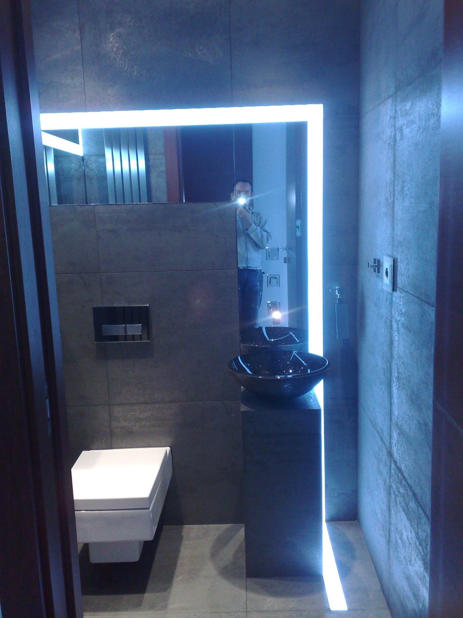 Nowoczesna łazienka z szarymi płytkami, podwieszaną toaletą, czarną umywalką na postumencie i lustrem podświetlanym LED, z widocznym odbiciem osoby robiącej zdjęcie.