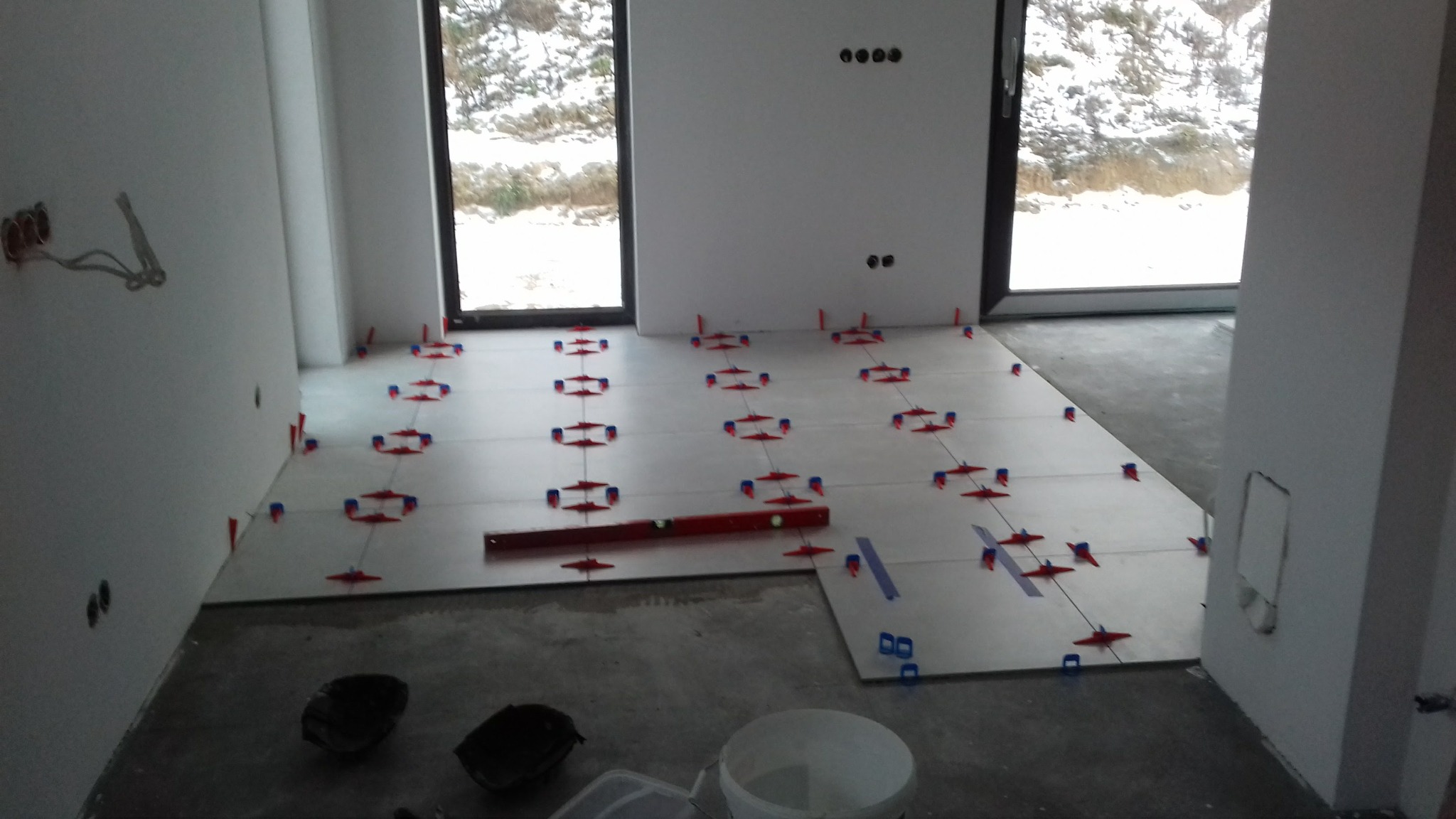 Układanie jasnych płytek podłogowych w pomieszczeniu z oknami, widoczne poziomica, krzyżyki dystansowe i przygotowane narzędzia.