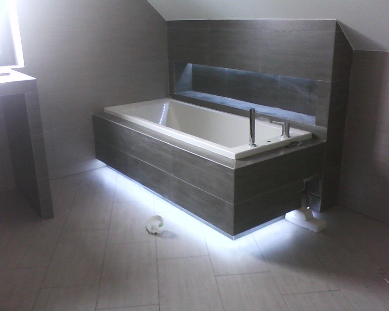 Nowoczesna wanna obudowana ciemnymi płytkami z podświetleniem LED, w tle ściana z wnęką również wyłożona płytkami, widoczne fragmenty armatury i niedokończone prace wykończeniowe.